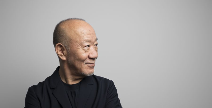 Composer Joe Hisaishi is in a black shirt and black suit. He has his arms folded and is looking away to the right of the photograph