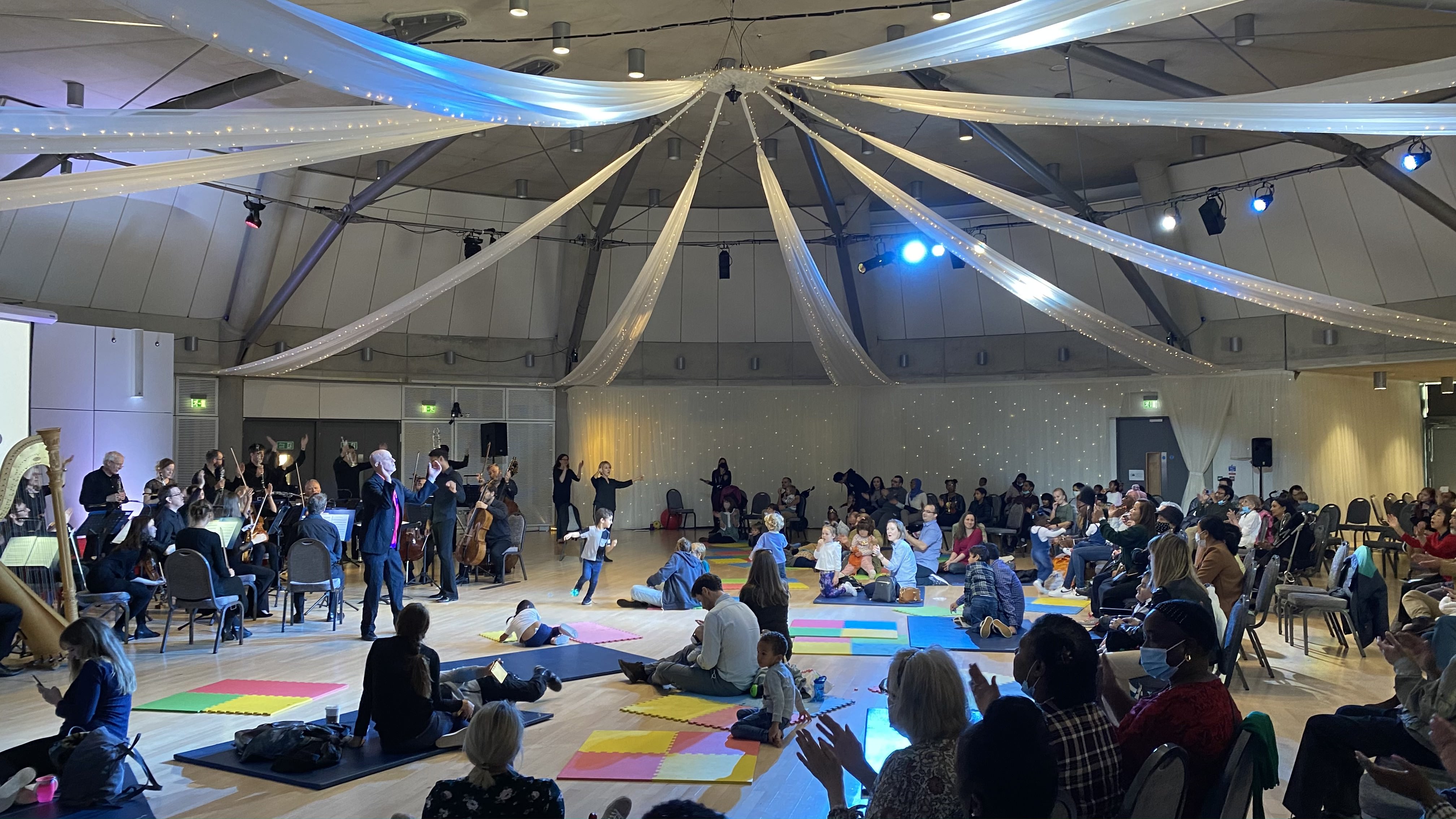 An image showing a small orchestra performing in the Brent Civic Centre Dome. There is a man who is the presenter talking to the audience. The audience are all looking at him. Some of the audience are sitting on chairs, some are on the floor on mats. The audience are young children and adults.