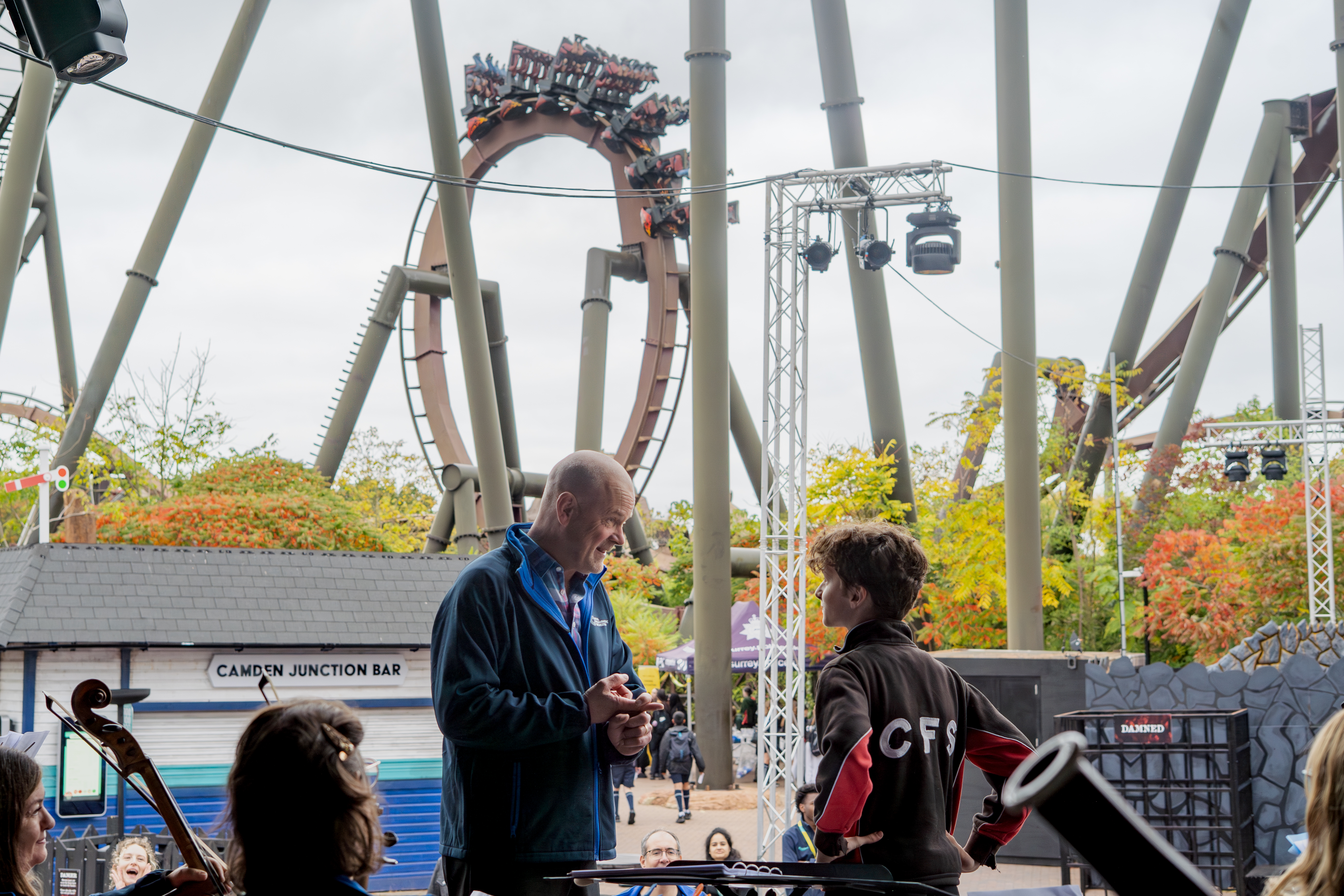Conductor, participant and Orchestra on stage with roller coaster in the background