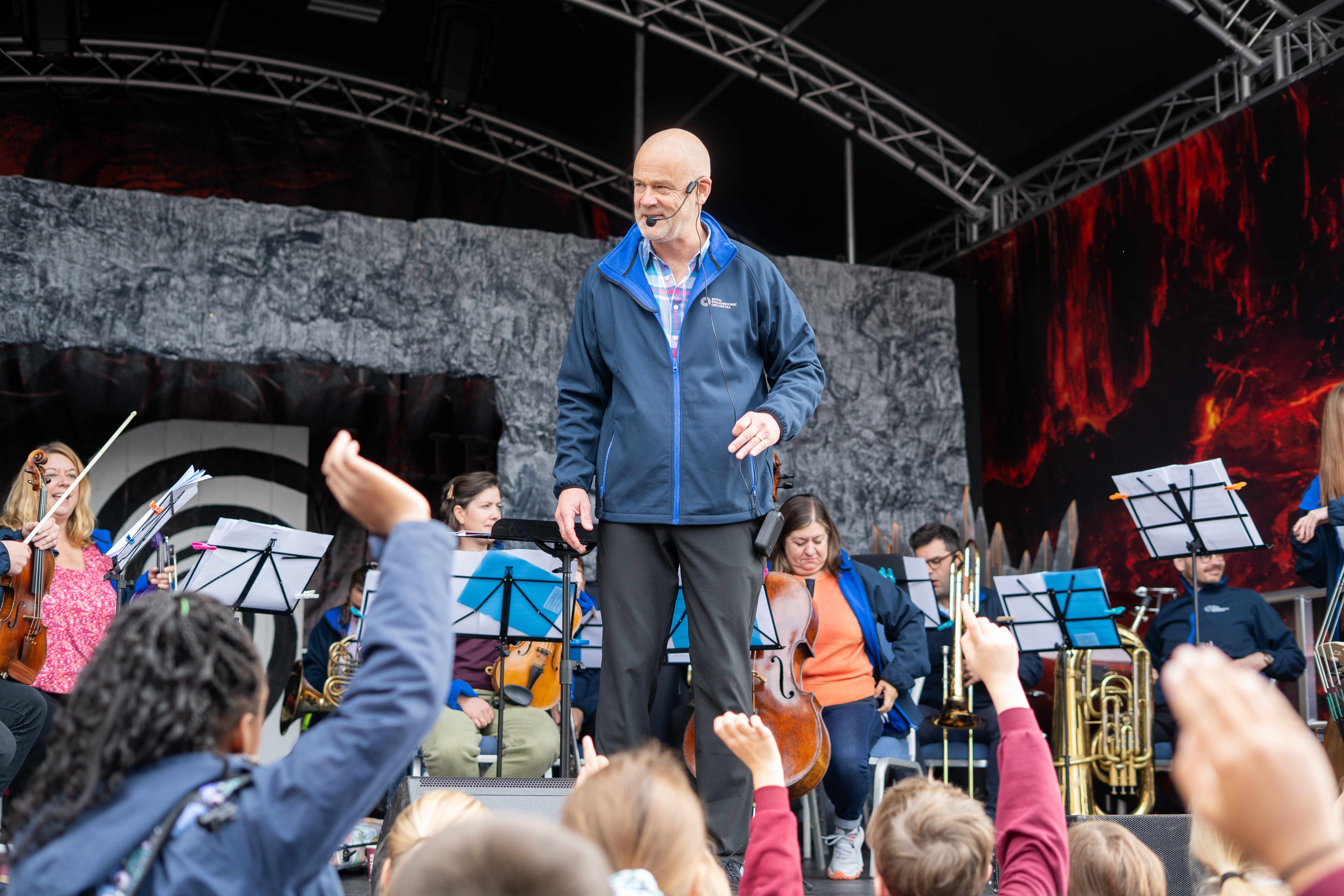 Conductor and Orchestra on stage, young people with hands raised to participate