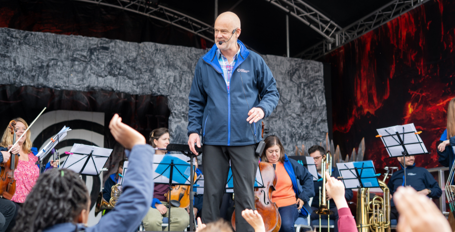 Conductor and Orchestra on stage, young people with hands raised to participate