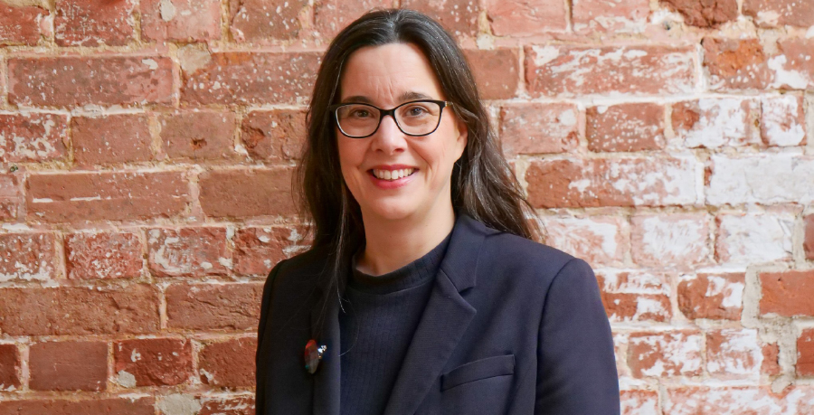 Sarah Bardwell is standing in a dark suit against a brick wall