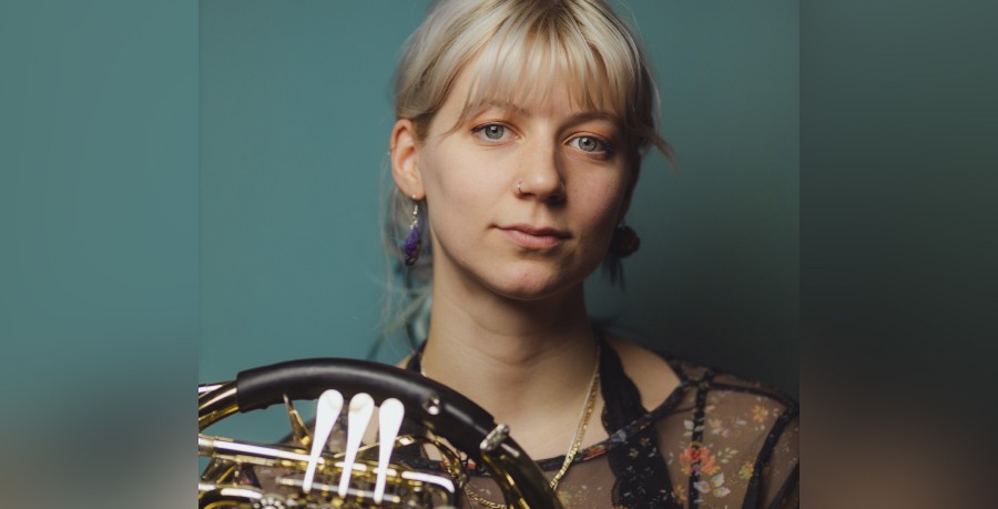 Horn player Zoë Tweed holding her horn and looking at the camera in front of a blue background