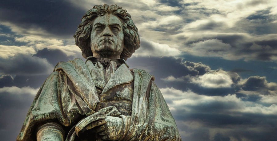 A statue of Beethoven with the blue-green colour of the copper patina process, against a dramatic sky with grey clouds.