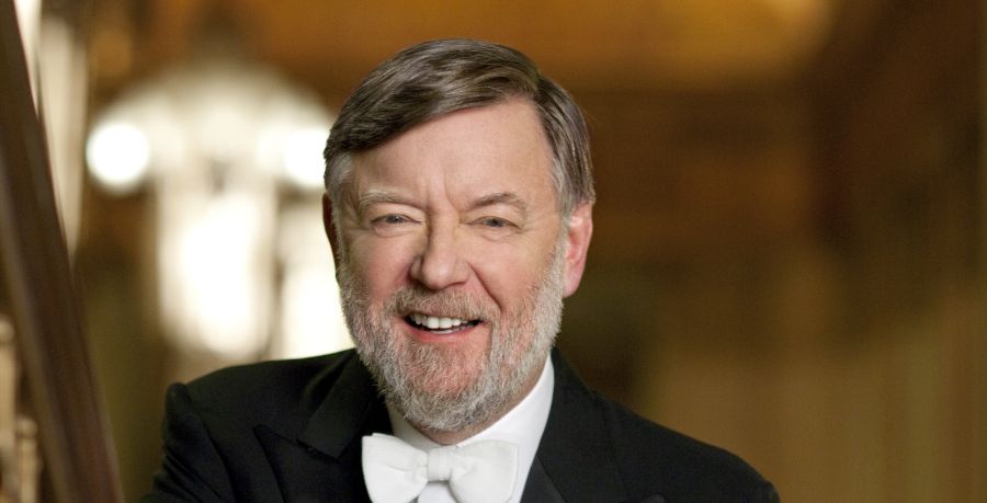 Conductor Sir Andrew Davis is wearing a white tie and tails. He is smiling leaning on a gold bannister looking straight at the camera.