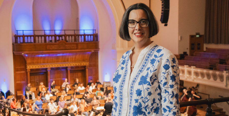 Sarah Bardwell in  white cardigan will blue flowers standing and smiling on the upper Gallery at Cadogan Hall, with the Orchestra rehearsing behind her
