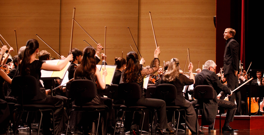 The first violins raising their bows in the air after playing a final note, and conductor Vasily Petrenko standing on the podium and smiling