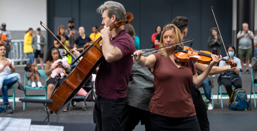 An RPO string quartet playing standing up and back to back in a car park in Wembley Park, with a crowd sitting and standing behind them