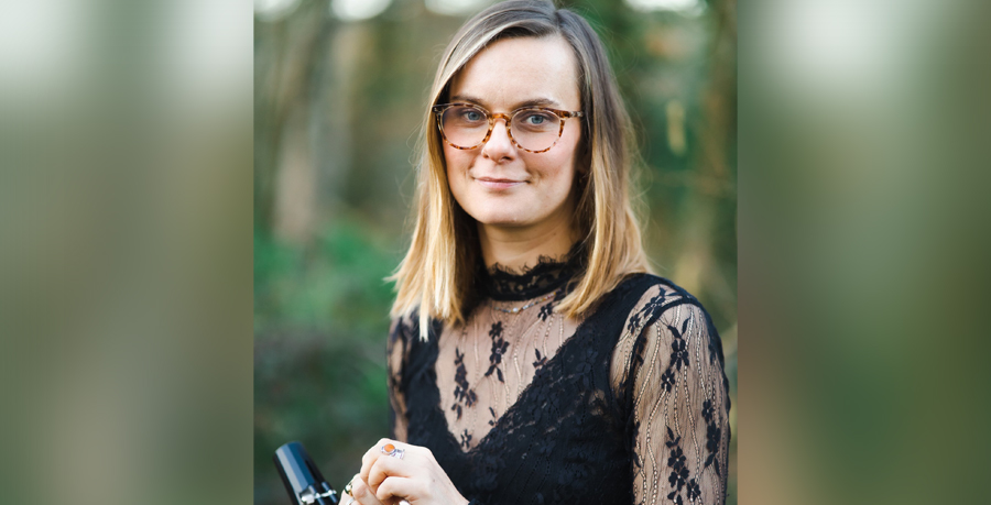 Katy Ayling in black concert dress holding her clarinet in front of some greenery