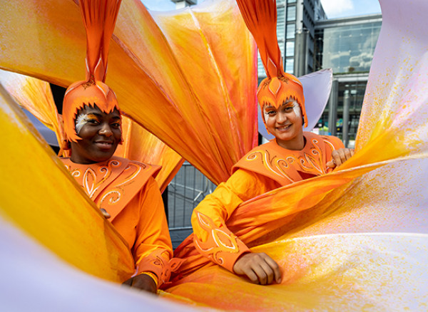 An image of two people wearing bright orange costumes. In the background is a glass building.