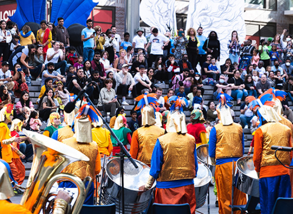 An image of standing drummers performing outside with an audience sitting on steps watching. The drummers are wearing a 