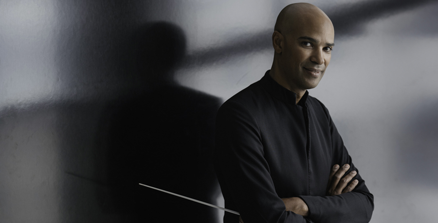 Conductor Kevin John Edusei holding a baton and leaning against a reflective wall