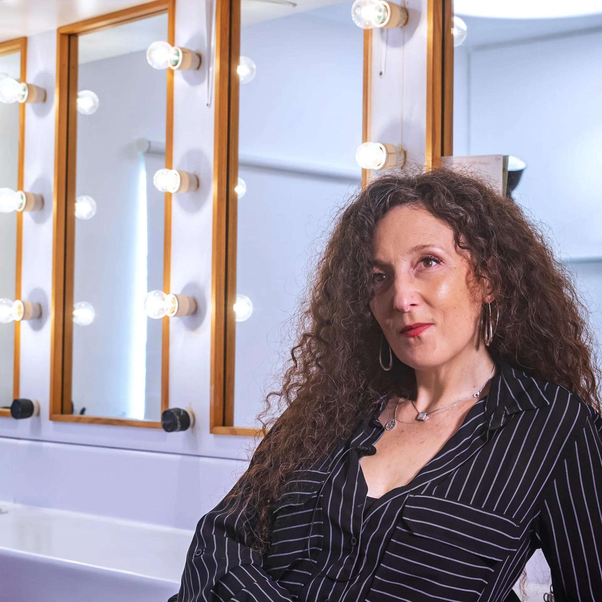 Nora Gubisch is on the right hand side of the photo. She is in a dressing room with a dressing table and mirrors with lightbulbs around them behind her.