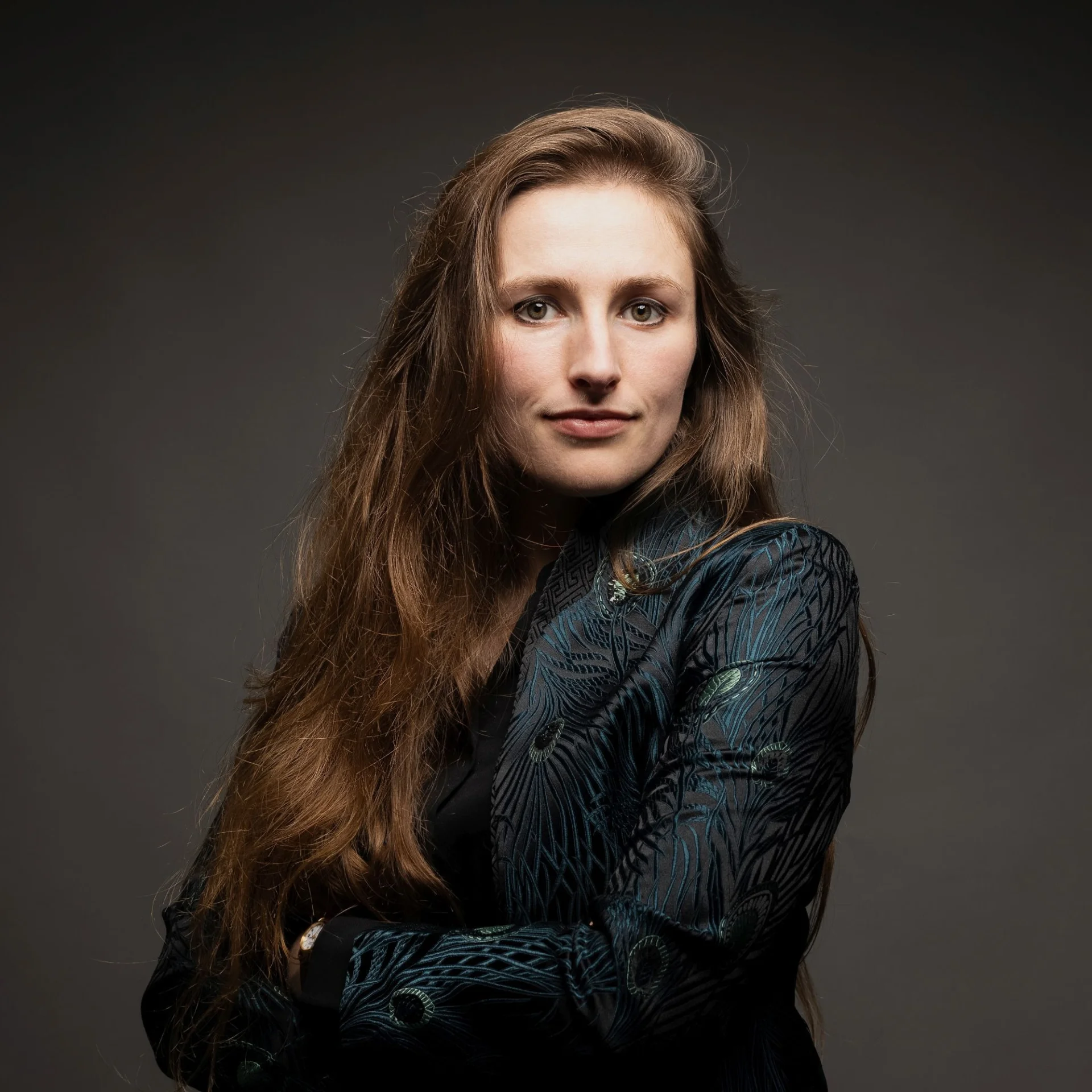 Leah Broad is shown in a dark jacket with peacock feathers depicted on the jacket with long hair over one shoulder, looking at the camera. She is against a dark background.