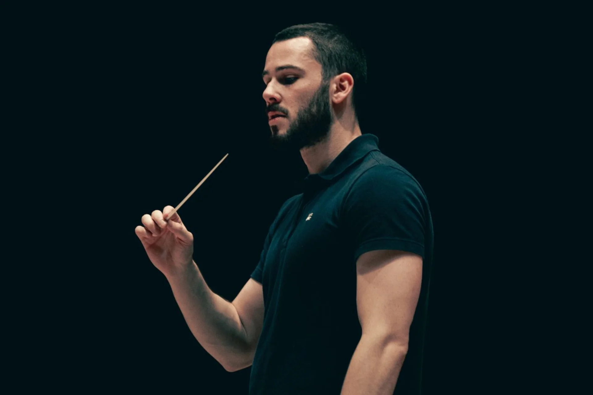 Conductor Oliver Zeffman stands with baton in hand, wearing a black polo shirt