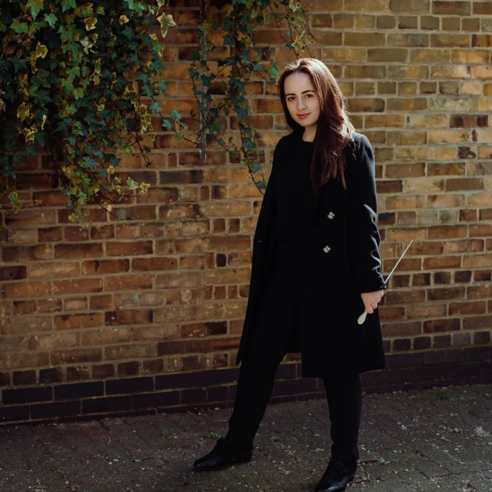 Conductor Olivia Clarke stand with baton in hand, looking into the camera