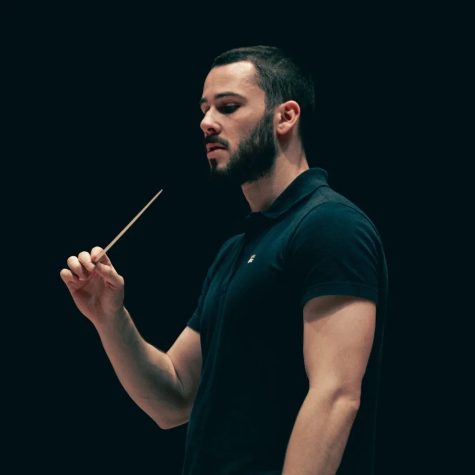 Conductor Oliver Zeffman stands with baton in hand, wearing a black polo shirt