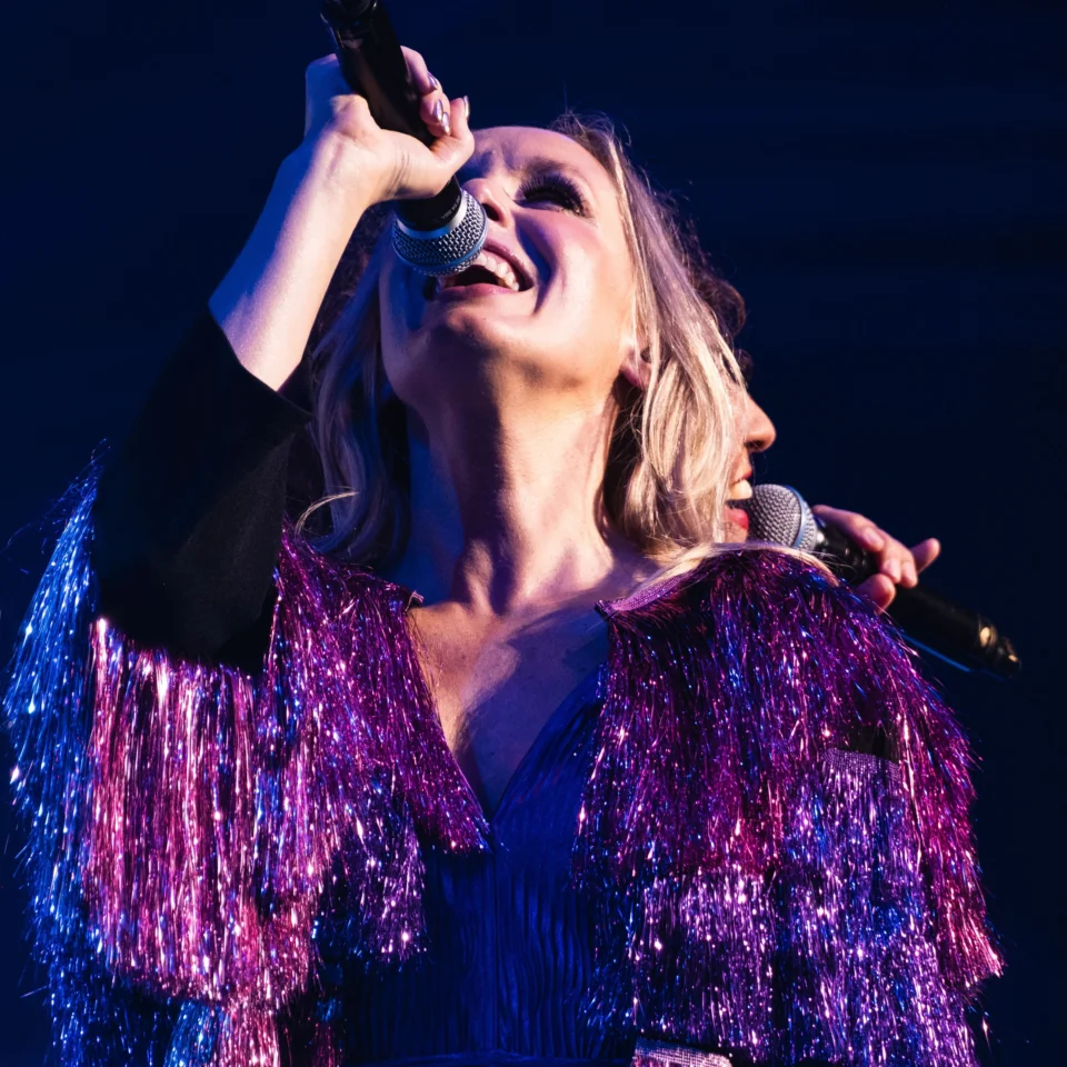Two vocalists stand back to back singing, wearing sparkly purple jackets