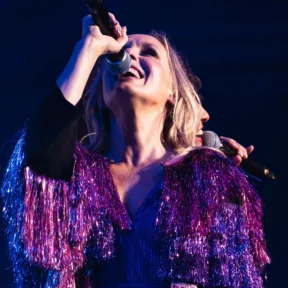 Two vocalists stand back to back singing, wearing sparkly purple jackets