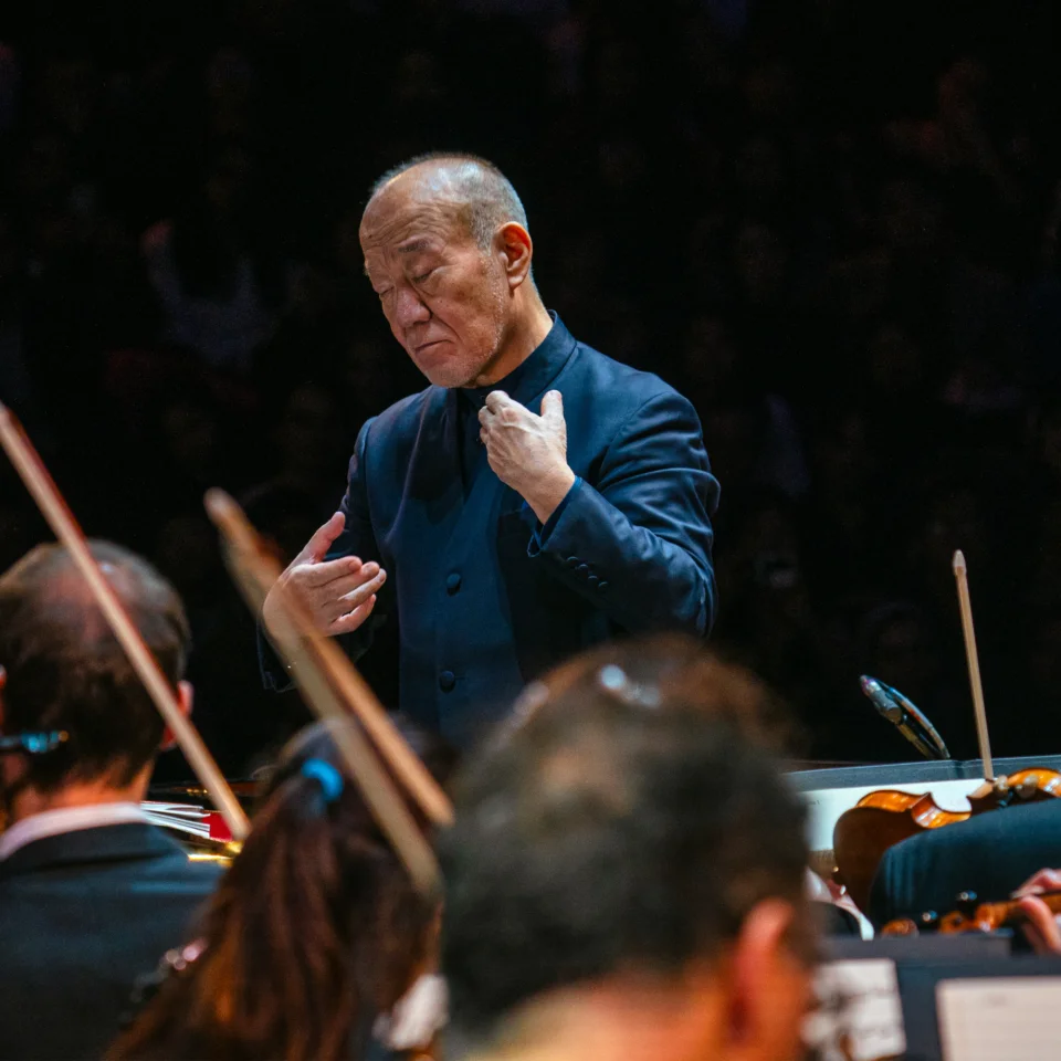 Joe Hisaishi conducting the Orchestra. His hands and held towards himself and his eyes are closed
