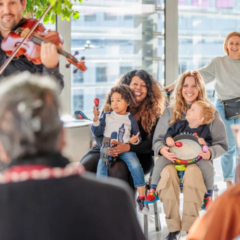 Round children with their parents sitting and playing percussion in a circle with an RPO violist playing near them