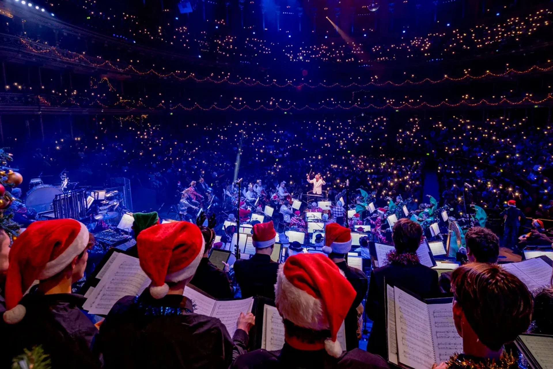 Choir singers in Christmas hats behind the Orchestra in the Royal Albert Hall. Audience members in the auditorium are holding up the torches on their phones