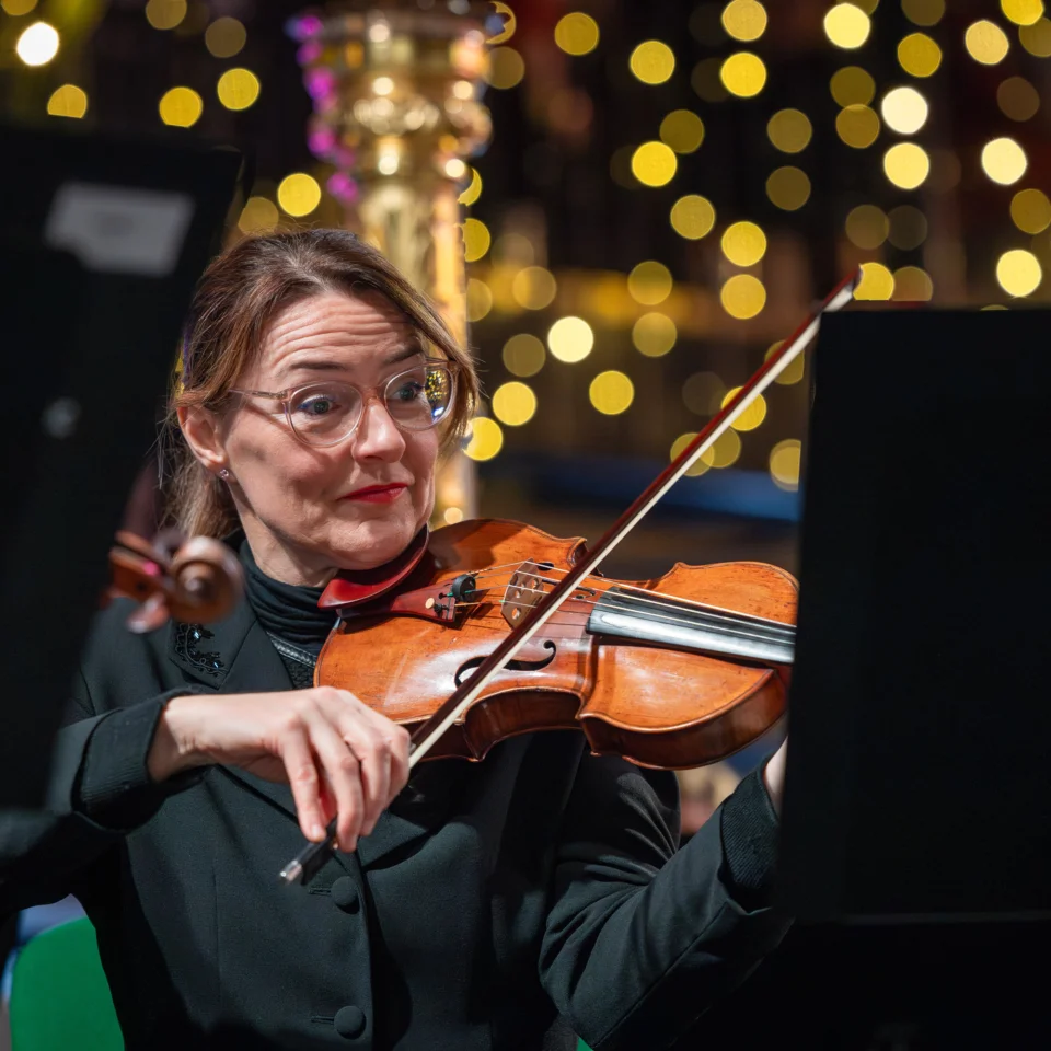 An RPO violinist playing on stage with a harp and sparkly Christmas lights behind her