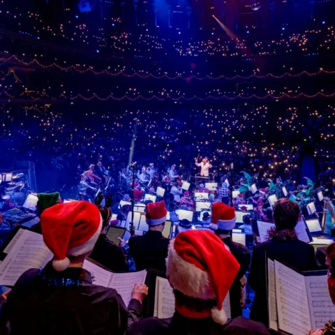 Choir singers in Christmas hats behind the Orchestra in the Royal Albert Hall. Audience members in the auditorium are holding up the torches on their phones