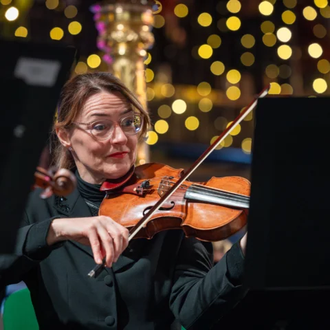 An RPO violinist playing on stage with a harp and sparkly Christmas lights behind her