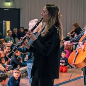 Royal Philharmonic Orchestra musicians play their instruments at a relaxed performance, integrating with the audience, some of which are sitting on floor mats