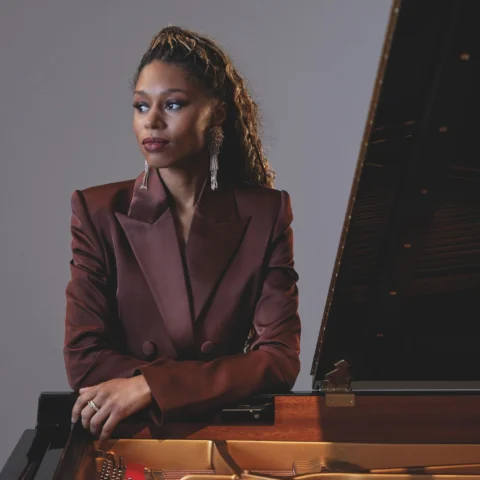 Isata Kanneh Mason stands leaning on a piano