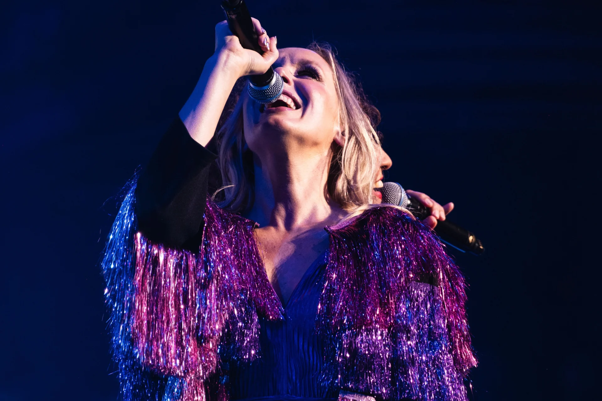 Two vocalists stand back to back singing, wearing sparkly purple jackets
