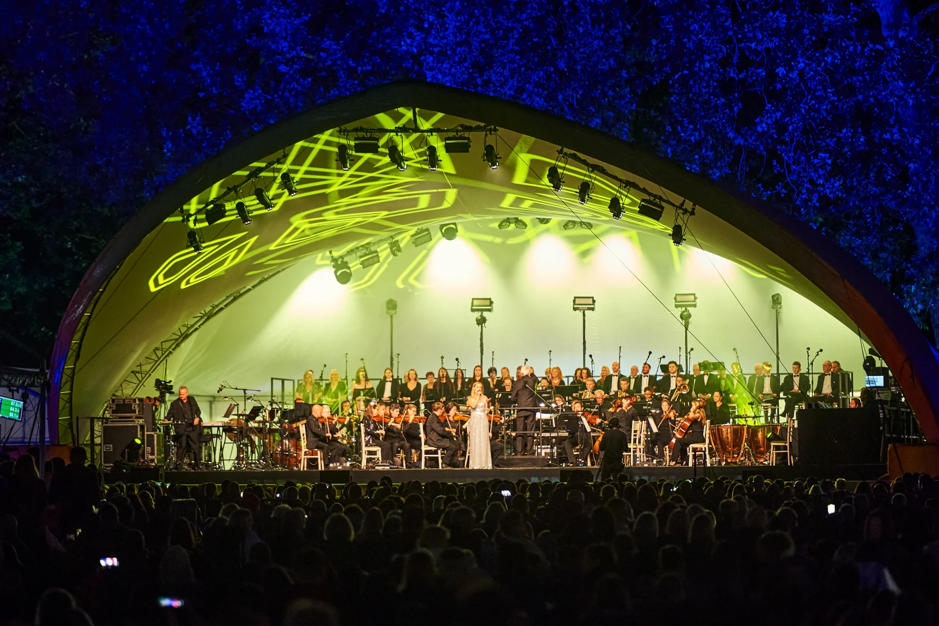 The RPCO performing on stage with a vocalist in front of the Orchestra in the evening at Battersea Park
