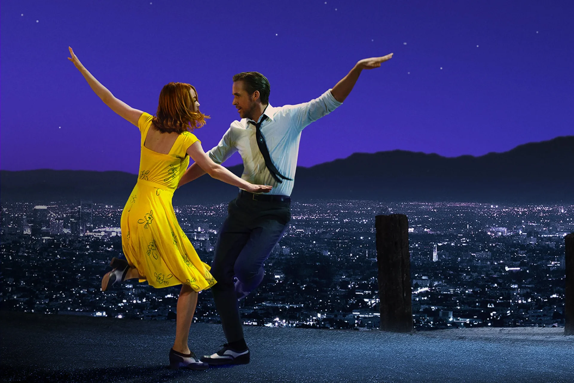 Two dancers in front of the LA skyline, one woman in a yellow dress and one man in a suit.