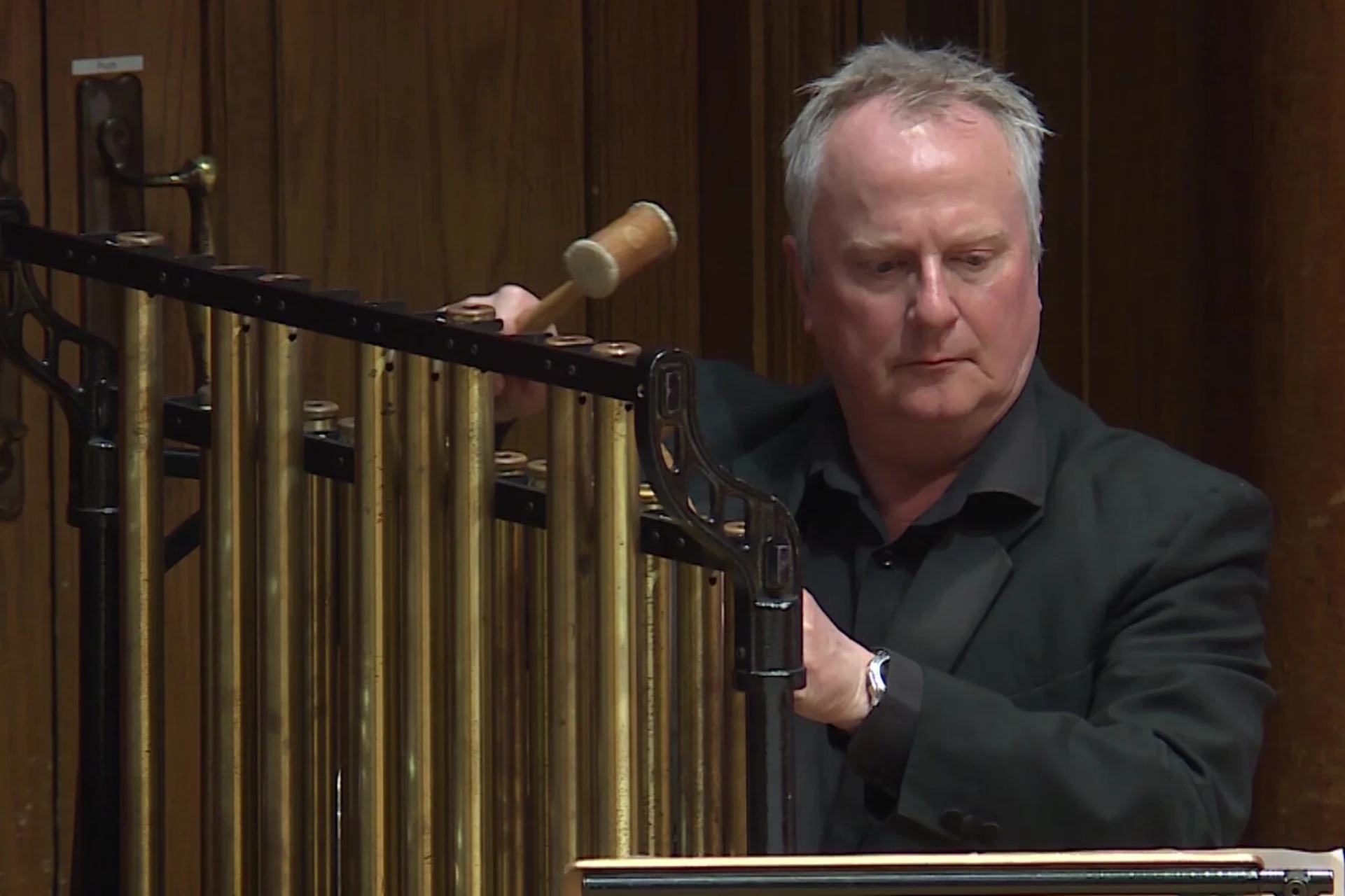 Gerald Kirby plays tubular bells in black shirt and jacket