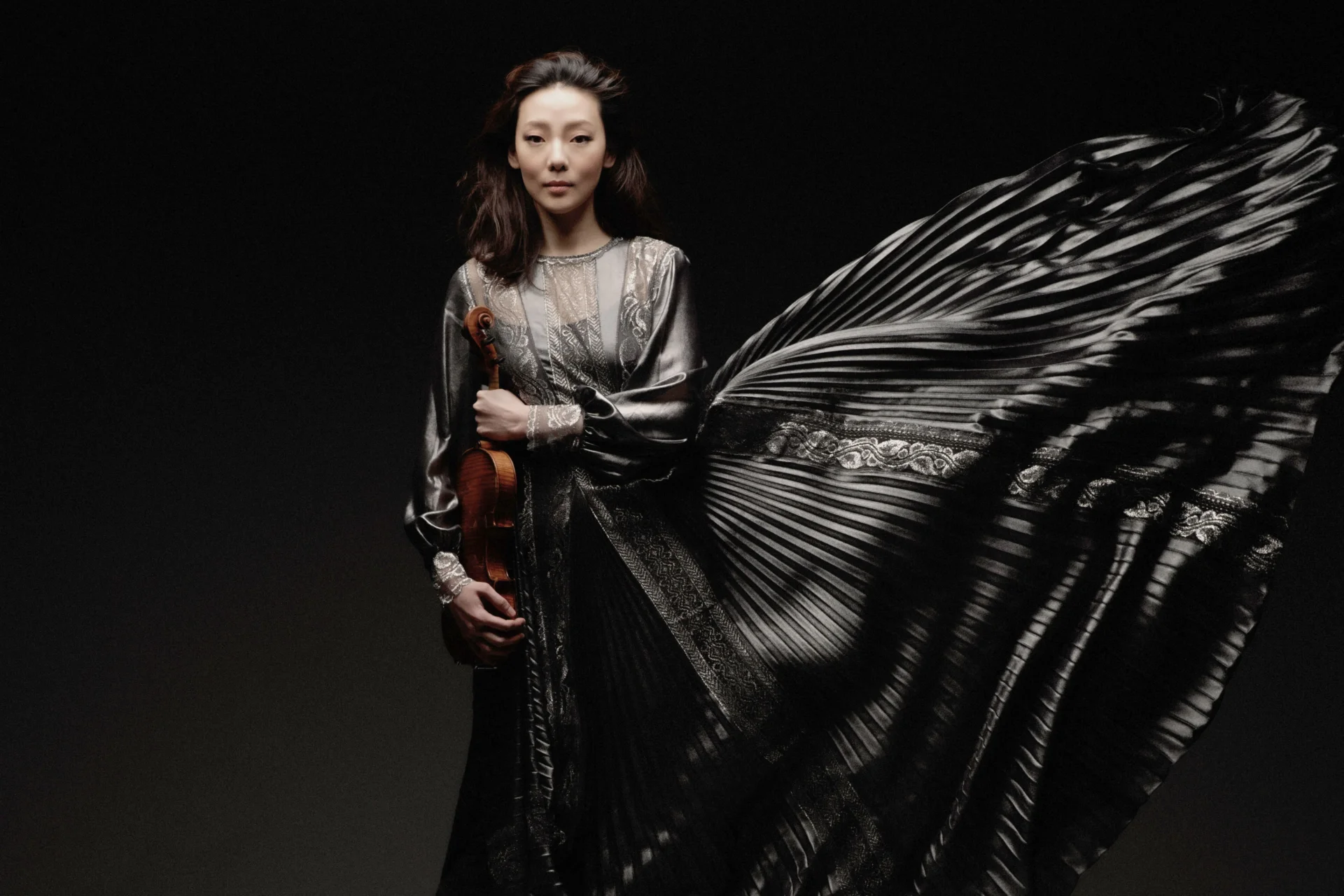 Violinist Clara Jumi Kang stands holding her violin in a long ruffled grey dress