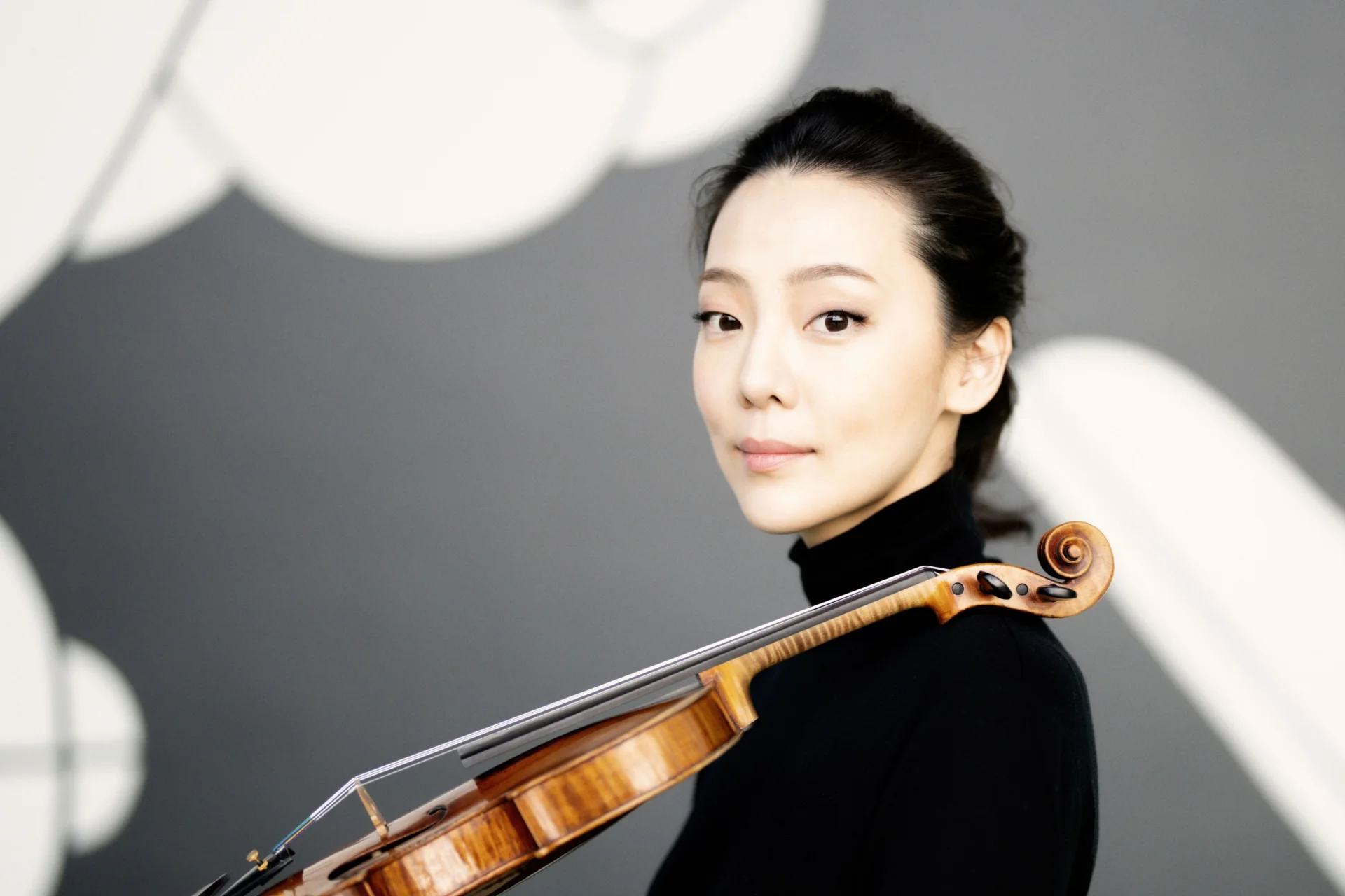 Clara Jumi Kang stands with her violin resting on her shoulder, looking at the camera.