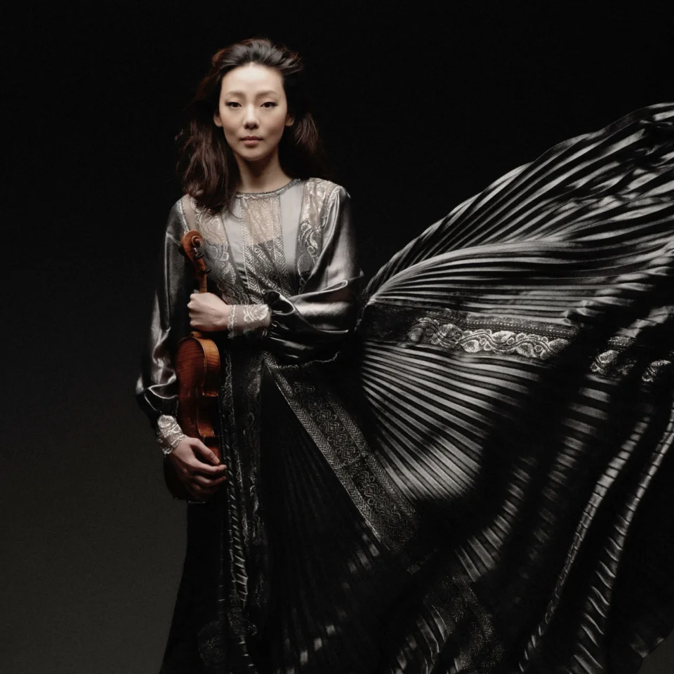 Violinist Clara Jumi Kang stands holding her violin in a long ruffled grey dress