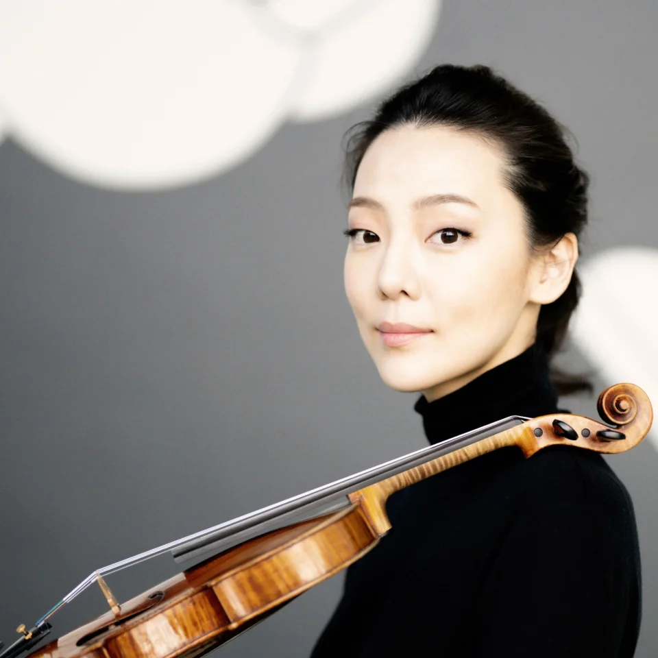 Clara Jumi Kang stands with her violin resting on her shoulder, looking at the camera.