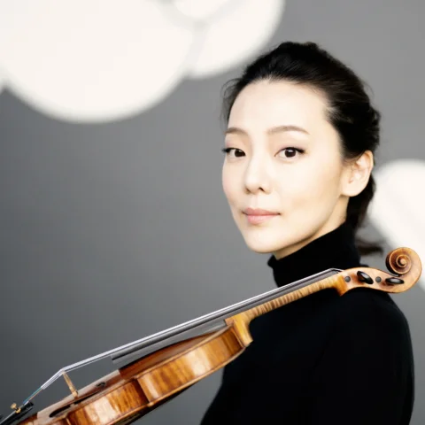 Clara Jumi Kang stands with her violin resting on her shoulder, looking at the camera.