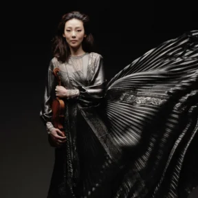 Violinist Clara Jumi Kang stands holding her violin in a long ruffled grey dress
