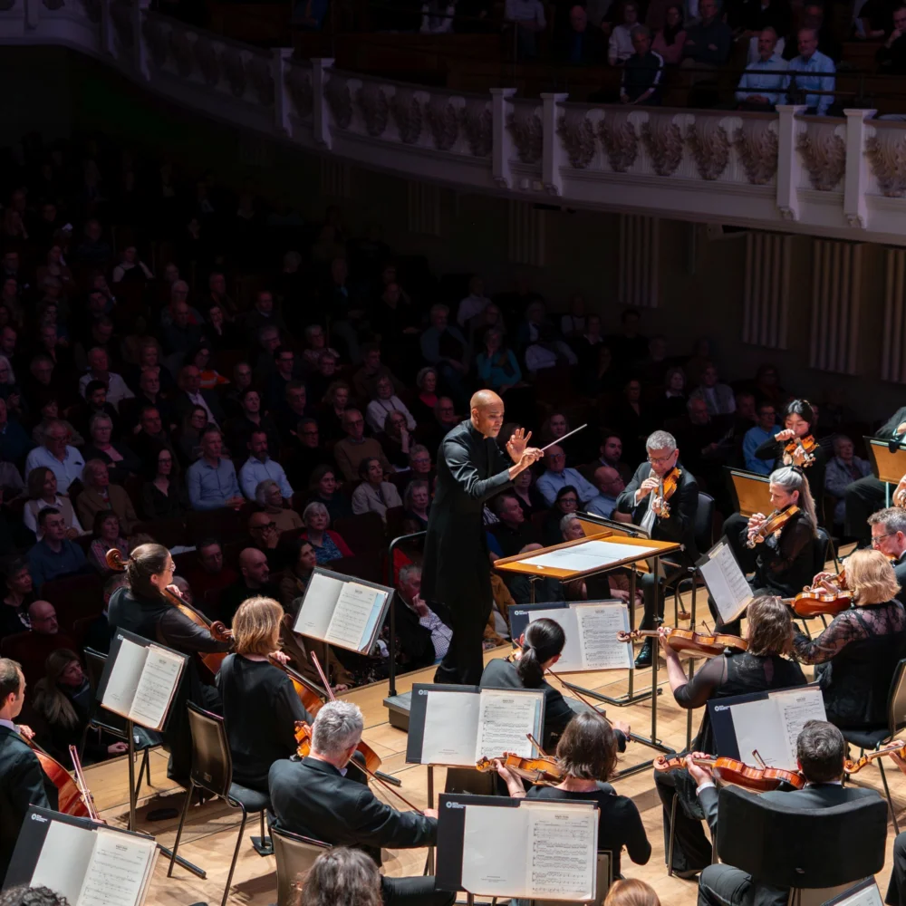 A photo taken from behind the Orchestra looking out into Cadogan Hall with conductor Kevin John Edusei shown at the front of the stage, slightly crouched with both hands in front of him, looking out at the musicians