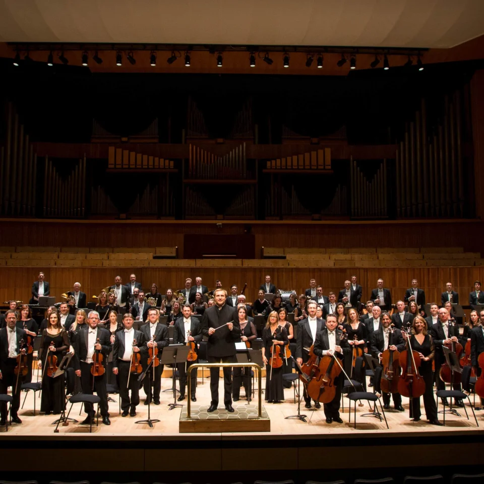 All of the Royal Philharmonic Orchestra stands on stage with their instruments, with conductor Vasily Petrenko in the middle