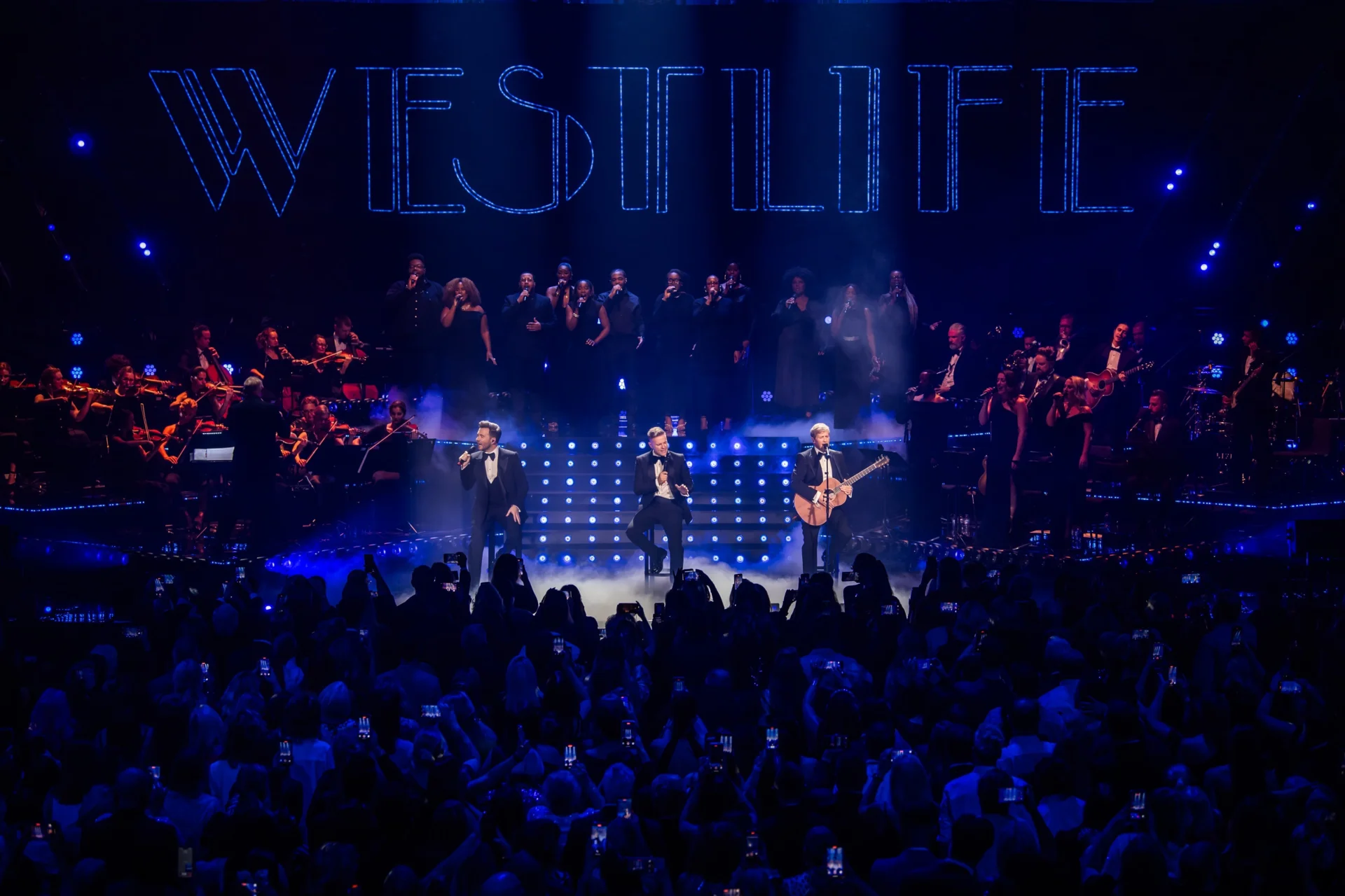 Three members of Westlife performing on stage at the Royal Albert Hall with the RPCO, singers and band to the sides