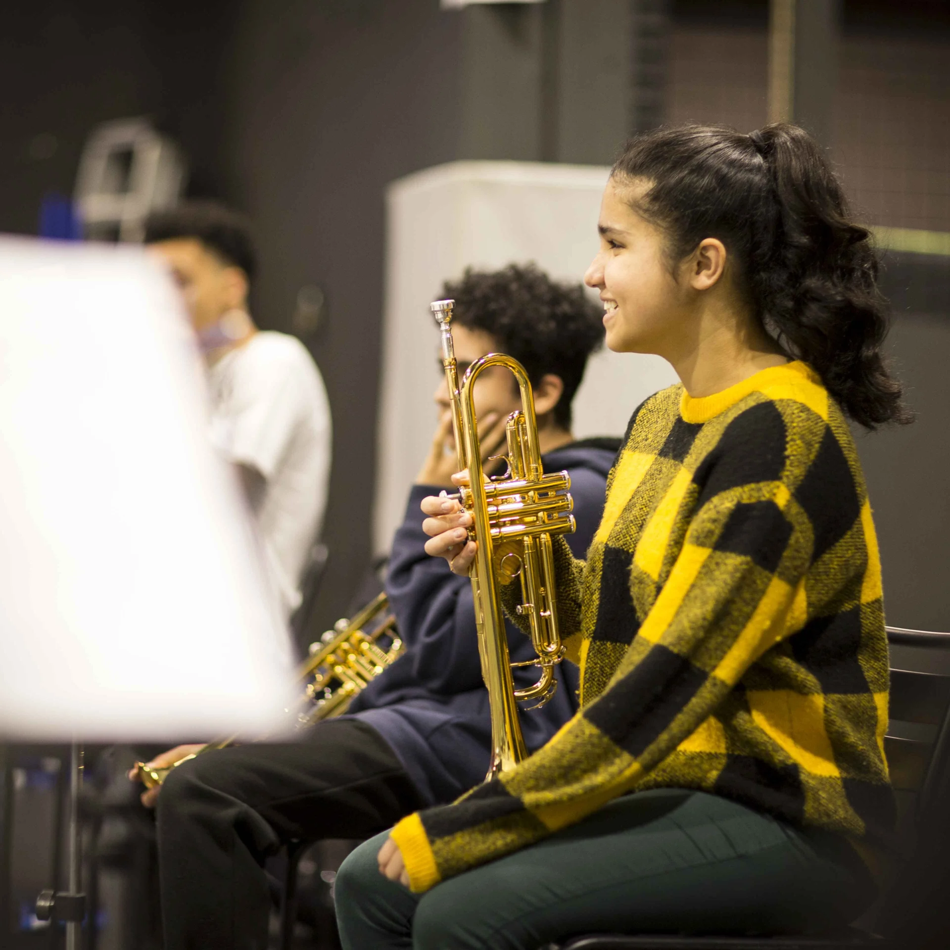A girl in a yellow and black jumper sits with her trumpet on her knee. She is smiling.