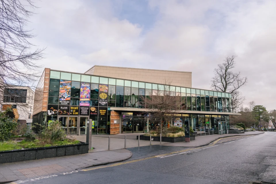 Exterior shot of the venue G Live in Guildford on a bright day. The building features a largely glass exterior and surrounding trees.