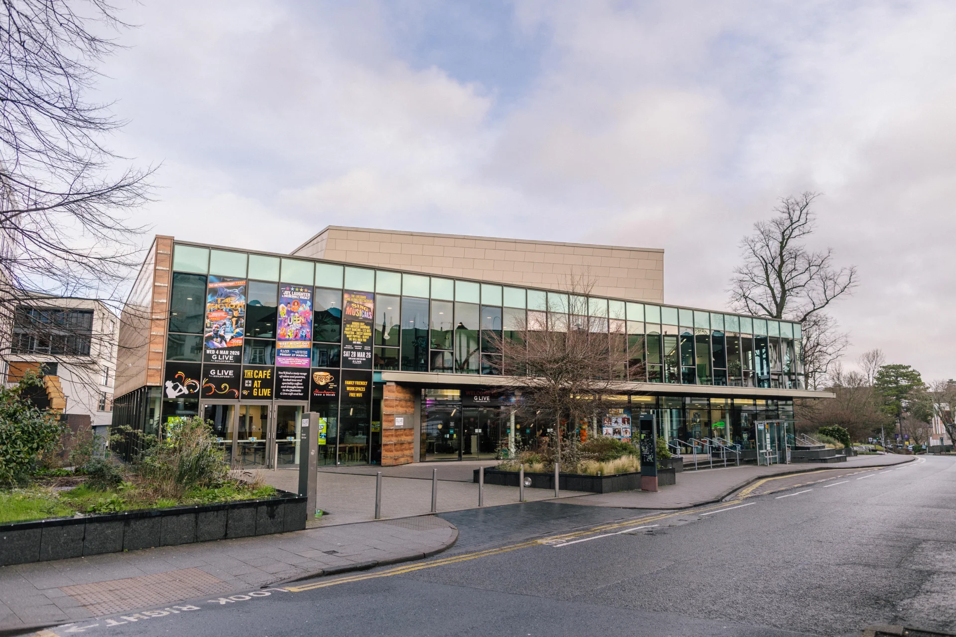 Exterior shot of the venue G Live in Guildford on a bright day. The building features a largely glass exterior and surrounding trees.