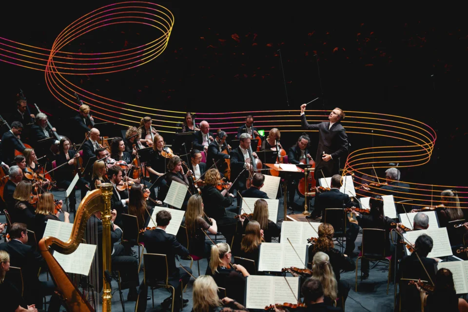 Vasily Petrenko conducting the RPO at the Roya Albert Hall with colourful musical stave lines winding behind him and the orchestra