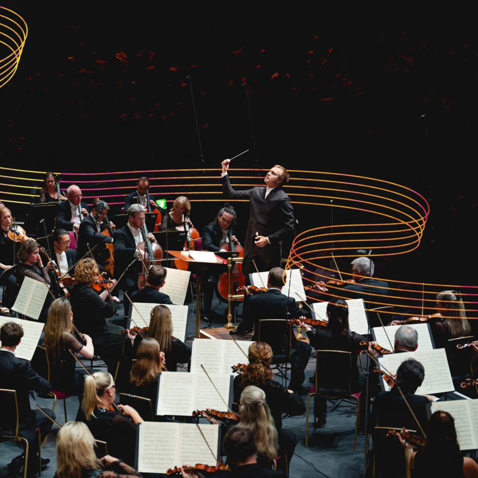 Vasily Petrenko conducting the RPO at the Roya Albert Hall with colourful musical stave lines winding behind him and the orchestra