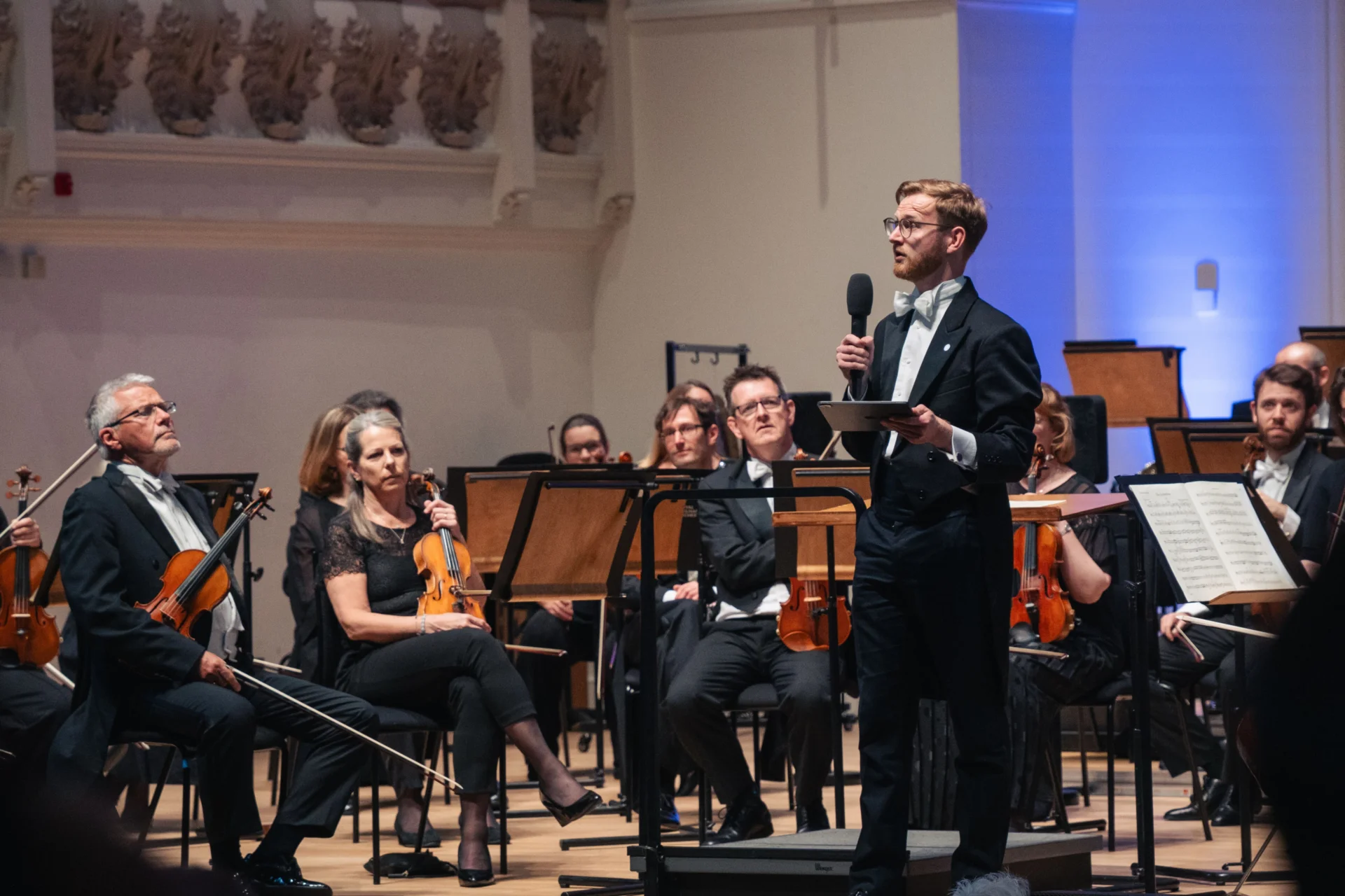 Double Bass player Joe Cowie stands on stage at Cadogan Hall with the Orchestra, speaking to the audience about the repertoire